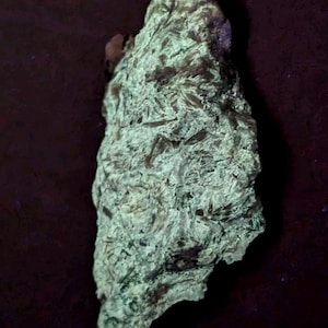 May include: A rough, irregularly shaped rock formation with a light green and brown color palette. The surface has a textured, layered appearance, with visible crystalline structures. The rock is set against a dark background.
