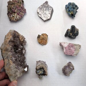 May include: A collection of eight distinct mineral specimens including a large purple amethyst geode, a metallic silver mica sheet, a cluster of blue crystals, a golden pyrite formation, and various other raw rock and crystal formations of different shapes.