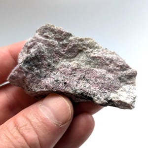 May include: A hand holds a rough, irregularly shaped rock. The rock has a mix of colors, including pink, gray, and black. The texture appears coarse and uneven, with visible mineral grains. The background is plain white.