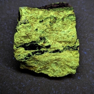 May include: A close-up of a mineral specimen with a vibrant, neon-yellow hue. The rock has a rough, textured surface with dark black veins running through it. The specimen is set against a dark, textured background.