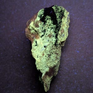 May include: A rough, irregularly shaped rock with a vibrant, neon green glow. The rock has a textured surface with dark black patches and a brown base. The image is taken against a dark purple background.