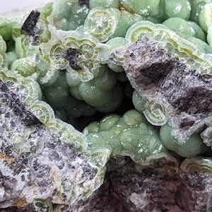 May include: A close-up of a green and black mineral specimen with a rough, textured surface. The mineral has a concentric, layered structure with a green color.
