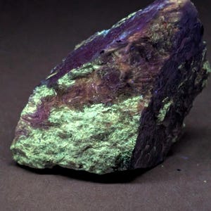 May include: A close-up of a mineral specimen with a rough, textured surface. The rock displays a mix of colors, including deep purple, light green, and brown. The lighting highlights the crystalline structure and variations in color.