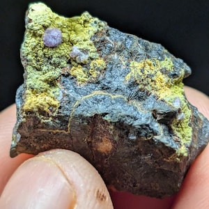May include: A close-up of a rough, dark gray rock with patches of yellow-green and a small, light purple crystal. The rock has a textured surface and is held in a person's hand. The background is black.