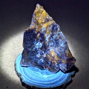 May include: A rough-cut mineral specimen with a mix of yellow, brown, and blue hues. The rock sits on a small, round wooden base with visible growth rings. The lighting highlights the mineral's texture and color variations.