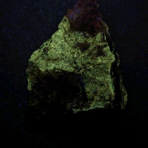 May include: A close-up of a rock formation with a rough, textured surface. The rock has a dark base with a vibrant green glow, and a reddish-brown top. The image is set against a dark background.
