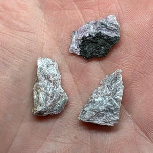 May include: Three rough-cut mineral specimens with a layered, crystalline structure. The stones display shades of gray, white, and a hint of purple. One piece has a dark green inclusion. The minerals are held in a hand.