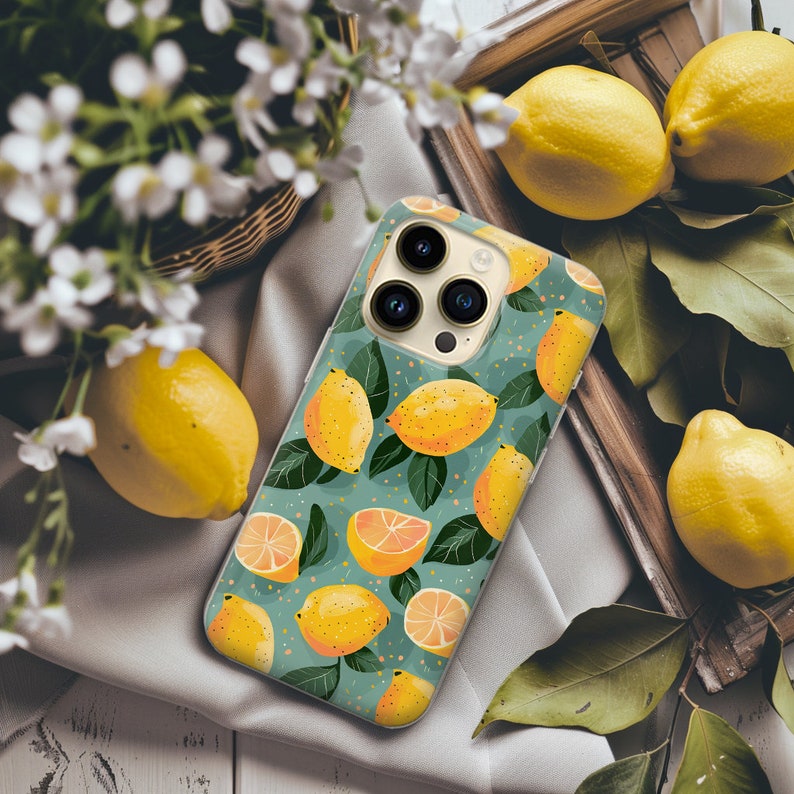 May include: A green, white, and yellow phone case with a lemon pattern and a camera lens design. The case is on a white fabric surface with lemons.