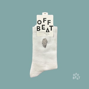 Könnte beinhalten: Weiße gerippte Socken mit einer Austernstickerei. Die Socken haben ein Etikett mit dem Text "OFF BEAT" in einer modernen Schriftart. Der Hintergrund ist türkis.