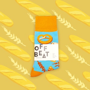 Op de afbeelding: Een paar blauwe en oranje sokken met een pretzel-, brood- en tarweontwerp. De sokken hebben de tekst "OFF BEAT" aan de zijkant.