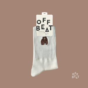 Könnte beinhalten: Eine weiße, gerippte Socke mit einem kleinen, braun gestickten Bären im oberen Bereich. Die Socke ist an einem weißen Pappetikett befestigt, auf dem oben der schwarze Schriftzug OFF BEAT und darunter weiterer kleiner Text zu sehen ist.