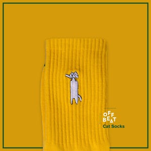 Może przedstawiać: Żółte skarpetki bawełniane z haftowanym białym wzorem kota. Na skarpetkach widnieje napis "OFF BEAT" i "Cat Socks".