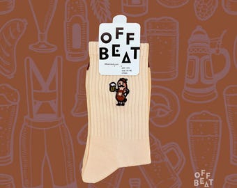 Pode incluir: Meias bege com uma imagem de desenho animado de um homem segurando uma caneca de cerveja. As meias têm uma textura canelada e uma etiqueta que diz "OFF BEAT". O fundo apresenta ilustrações relacionadas à cerveja.
