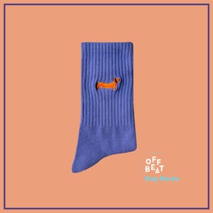 Könnte beinhalten: Lilafarbene Socken mit einem aufgestickten orangefarbenen Dackel auf der Vorderseite. Die Socken tragen den Schriftzug "Off Beat Dog Socks" auf der Unterseite.