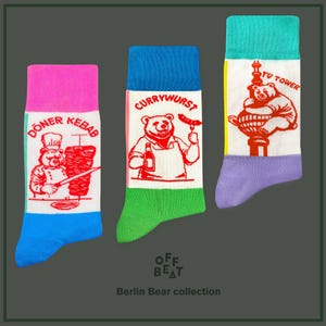 Puede incluir: Tres pares de calcetines coloridos con temática de Berlín. Cada calcetín presenta una ilustración diferente de un oso con un alimento o un hito local. Los calcetines son azules, verdes y rosas con detalles blancos. Las ilustraciones son de un oso comiendo un Döner Kebab, un oso comiendo una Currywurst y un oso de pie en la cima de la Torre de Televisión.