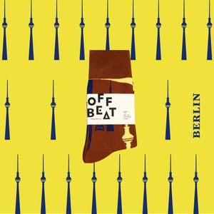 May include: Brown and yellow socks featuring a graphic print of the Berlin TV Tower. The socks come with packaging that reads "OFF BEAT". The background is yellow with a pattern of blue Berlin TV Towers.