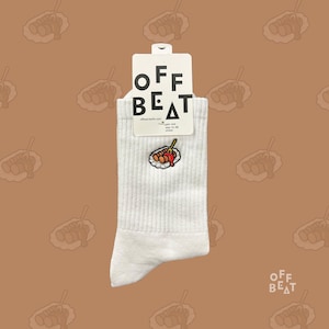 Op de afbeelding: Witte geribbelde sokken met een geborduurd beeld van een worst met ketchup op een bord. De sokken hebben een label met de tekst "OFF BEAT" in zwarte hoofdletters. De achtergrond is lichtbruin met een herhaald patroon van een worst.