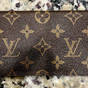 May include: A rectangular wallet featuring the iconic brown and tan monogram pattern with repeating LV initials and floral motifs. The exterior surface has a textured finish, and the design is consistent across the entire front of the item.