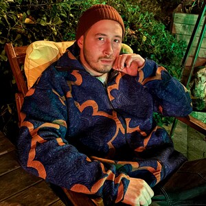 May include: A person wearing a brown beanie and a navy blue jacket with an orange floral pattern. The jacket is zipped up and the person is sitting in a wooden chair.