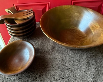 May include: A collection of wooden salad bowls and serving utensils. The set includes a large bowl, a smaller bowl, and a stack of individual bowls. Wooden salad servers rest on top of the stack. The bowls and servers are a warm brown color.