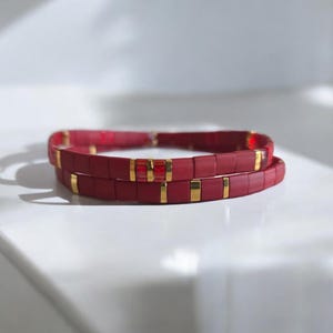 May include: Two red tile bracelets with gold accents. The bracelets are made of small, rectangular tiles strung together. Gold rectangular and square accents are interspersed throughout the bracelets. The bracelets are displayed on a white surface.