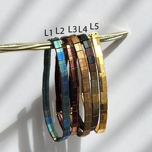 Könnte beinhalten: Fünf farbenfrohe Armbänder mit quadratischen Kacheln werden auf einem goldenen Stab präsentiert. Die Armbänder variieren in der Farbe, darunter Türkis, Blau, Bronze, Braun und Gold. Die Armbänder sind mit L1 bis L5 beschriftet.