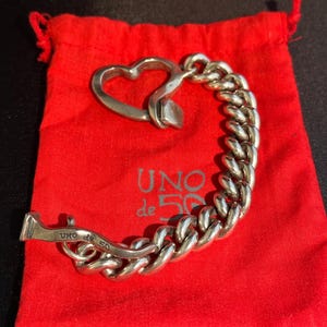 May include: A silver chain bracelet with a heart-shaped clasp. The bracelet is on a red velvet pouch with the words "UNO de 50" printed on it.
