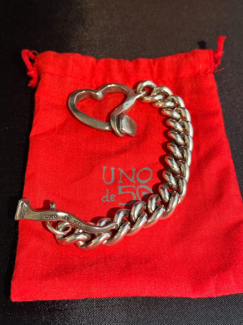 May include: A silver chain bracelet with a heart-shaped clasp. The bracelet is on a red velvet pouch with the words "UNO de 50" printed on it.