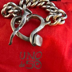 May include: A silver bracelet featuring a heart-shaped clasp and a chain link design. The clasp is shaped like a nail and has the words "UNO de 50" engraved on it.