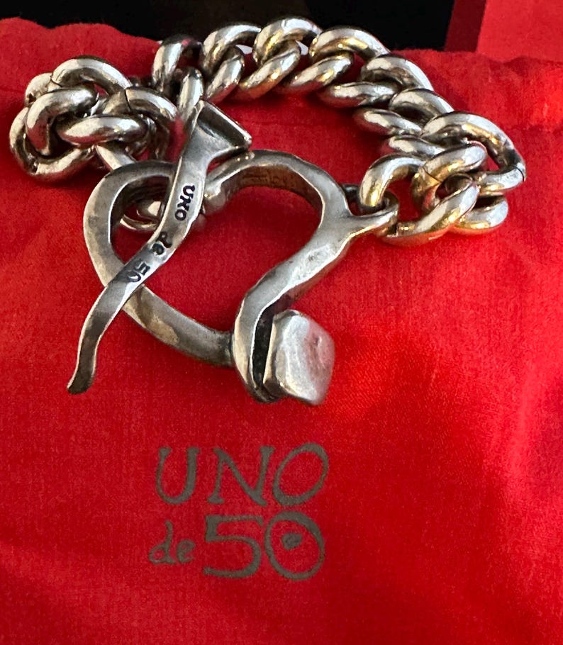 May include: A silver bracelet featuring a heart-shaped clasp and a chain link design. The clasp is shaped like a nail and has the words "UNO de 50" engraved on it.
