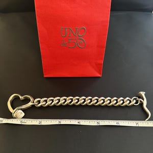 May include: A silver chain bracelet with a heart-shaped clasp and a hammer-shaped end. The bracelet is 17.8 cm long. The bracelet is on a white surface with a red bag with the text "UNO de 50" in gold lettering.