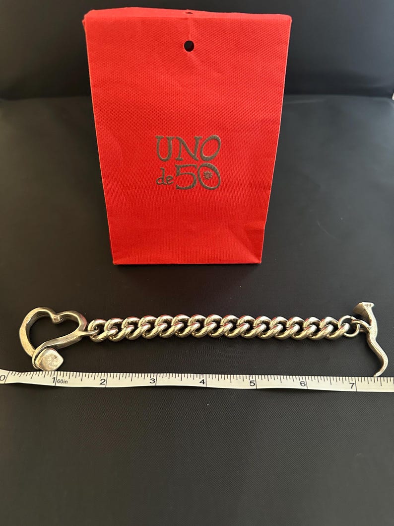 May include: A silver chain bracelet with a heart-shaped clasp and a hammer-shaped end. The bracelet is 17.8 cm long. The bracelet is on a white surface with a red bag with the text "UNO de 50" in gold lettering.