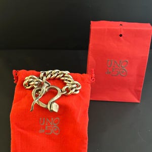 May include: A silver bracelet with a heart-shaped clasp. The bracelet is on a red velvet pouch with the words "UNO de 50" printed on it. The bracelet is also in a red paper bag with the same logo.