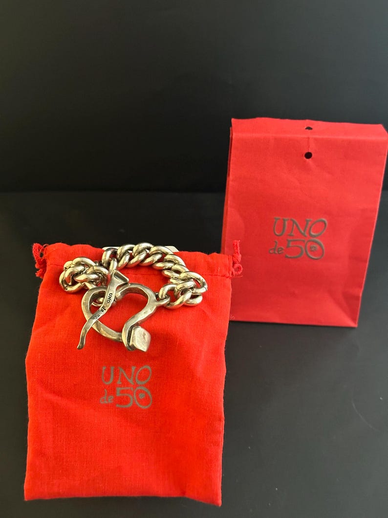 May include: A silver bracelet with a heart-shaped clasp. The bracelet is on a red velvet pouch with the words "UNO de 50" printed on it. The bracelet is also in a red paper bag with the same logo.