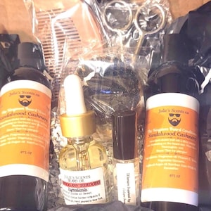 Puede incluir: Una caja de regalo con productos para el cuidado de la barba. Incluye dos botellas de acondicionador para barba, una botella de aceite para barba, un pequeño frasco de perfume, un peine y unas tijeras. Las botellas tienen etiquetas naranjas con texto.
