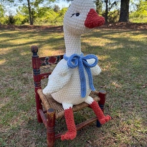 May include: A handmade, crocheted goose doll with a white body, red feet and beak, and a blue bow tie. The goose is seated on a small, antique wooden chair with a red frame and woven seat, set outdoors.