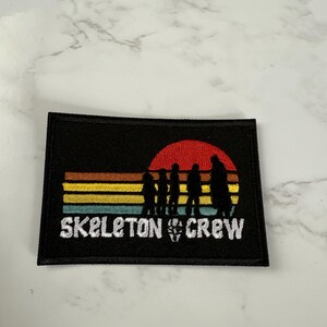 May include: Black rectangular patch featuring the words "SKELETON CREW" in white, with a skull graphic. The design includes a sunset with silhouetted figures and horizontal stripes in yellow, orange, and teal.