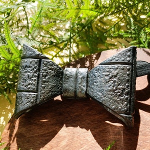 May include: A black bow tie with a textured, embossed surface. The bow tie has a subtle metallic sheen and is made of a durable material.