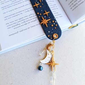 May include: A blue and gold star-themed bookmark with a dangling charm featuring a crescent moon, a star, and a blue bead. The bookmark is made of polymer clay and has a textured surface.