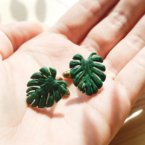 May include: Two green leaf-shaped cufflinks with gold-coloured posts. The cufflinks are designed to resemble Monstera plant leaves. The image is a close-up shot, highlighting the details of the leaves and the gold accents.
