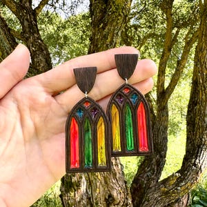 Può includere: Un paio di orecchini marrone scuro, in stile vetrata. Ogni orecchino presenta una parte superiore rettangolare e un design a vetrata con colori rosso, verde, giallo e blu. Gli orecchini sono sospesi da un gancio d'argento.