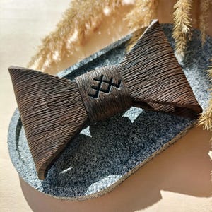 May include: A brown wooden bow tie with a carved design on the center. The bow tie is sitting on a gray stone coaster.