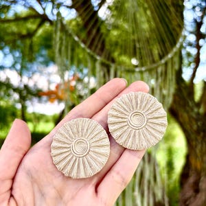Puede incluir: Dos pendientes circulares beige con un diseño de sol se sostienen en una mano. Los pendientes tienen un centro en espiral y flecos radiantes. El fondo está borroso, con follaje verde y un atrapasueños visible.