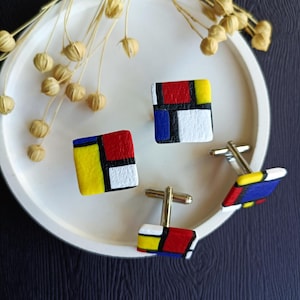 Op de afbeelding: Een set van drie manchetknopen met een geometrisch ontwerp geïnspireerd door het werk van Piet Mondrian. De manchetknopen zijn vierkant en hebben een zwarte omtrek met rode, gele en blauwe vierkanten erin. De manchetknopen liggen op een wit bord.