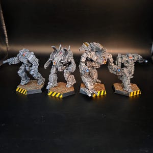 May include: Four miniature robot figures painted in shades of grey with metallic accents. Each figure stands on a hexagonal base featuring a yellow and black hazard stripe pattern along the edge and a textured brown surface on the top.