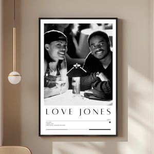 Puede incluir: Póster enmarcado en blanco y negro con el título "LOVE JONES" en letras blancas. La imagen muestra a una pareja sonriendo en una mesa. Debajo del título hay detalles adicionales. El póster está colgado en una pared beige.
