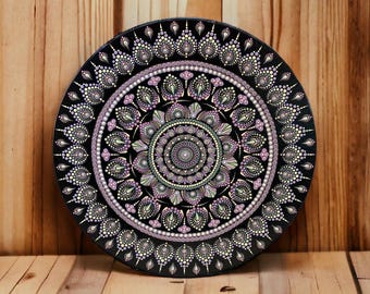 May include: A black, round decorative plate featuring a detailed mandala design. The mandala is composed of intricate patterns in shades of lavender, pale green, and cream-colored dots. The plate is displayed against a wooden background.
