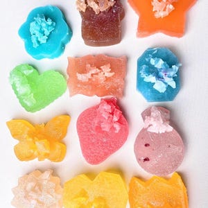 Puede incluir: Una colección de jabones coloridos con formas variadas, incluyendo una flor, un corazón, una estrella, una mariposa, entre otros. Cada jabón presenta un adorno cristalino en un color contrastante, creando una textura visual única. Los jabones están dispuestos sobre una superficie blanca.