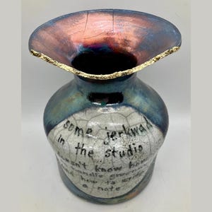 May include: A blue and white ceramic vase with a copper-colored rim and gold accents. The vase has a crackled glaze and features handwritten text that reads "some jerk wad in the studio doesn't know how to handle greenware and how it's with a note".