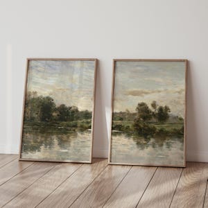 May include: Two vertical framed wall art prints featuring an impressionistic landscape painting of a calm river reflecting a line of dark green trees under a cloudy, muted sky with soft beige and grey tones, encased in light wood frames.
